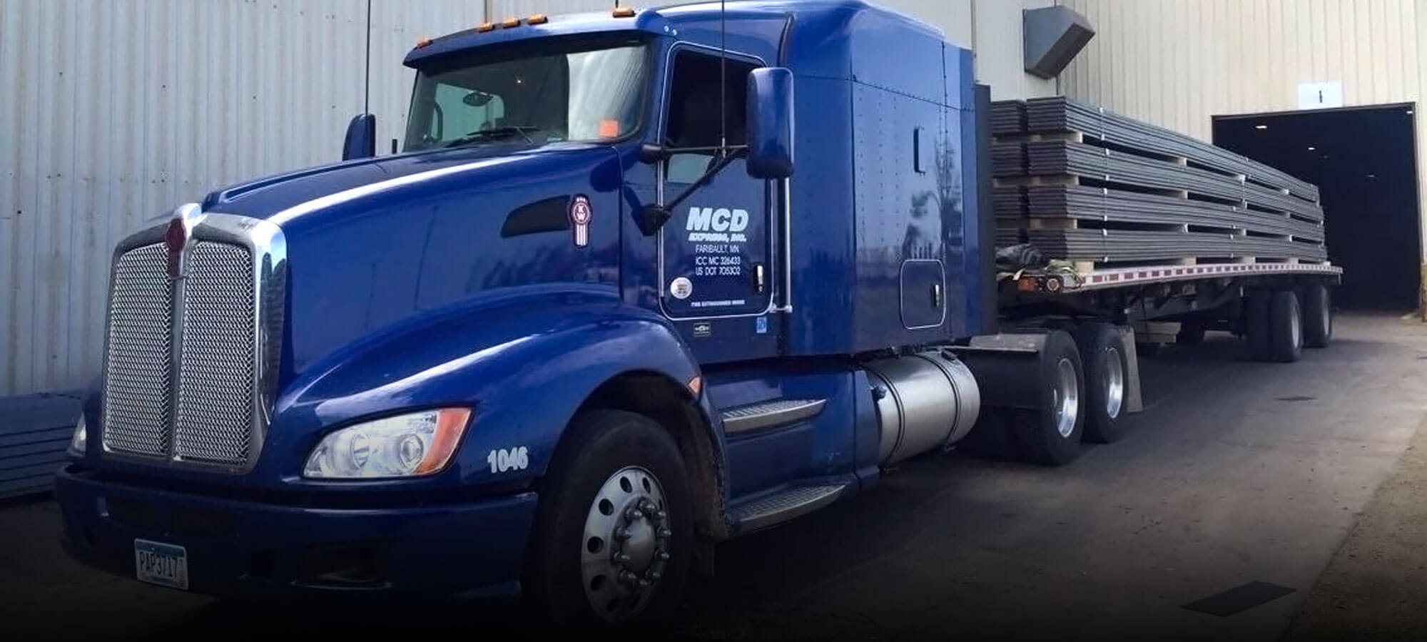 Royal blue MCD Express semi tractor hauling a flatbed trailer loaded with sheet metal